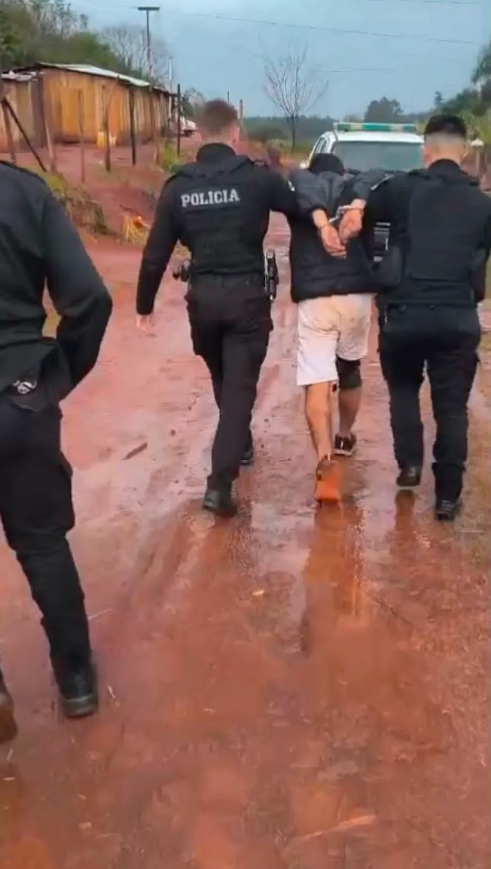 Dos policías llevan detenido y esposado a un hombre por un camino de tierra colorada.
