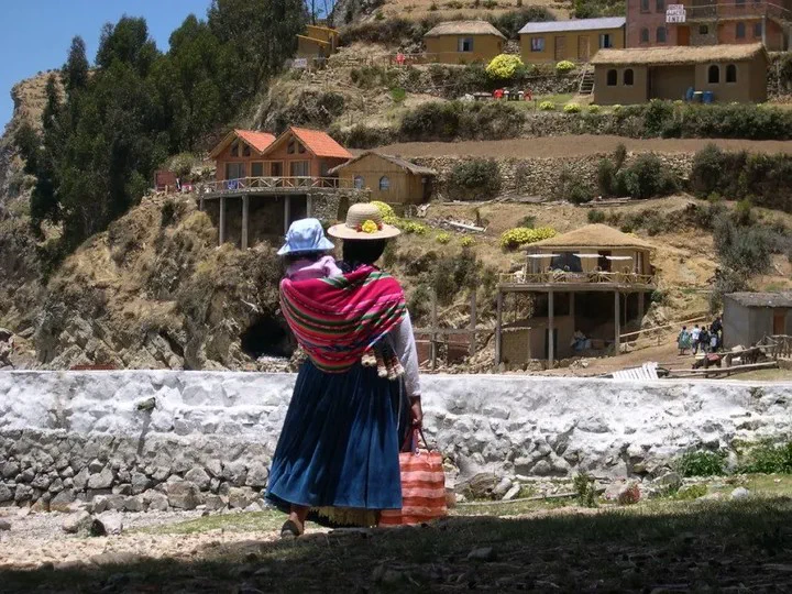 Una mujer con vestimenta tradicional andina y un sombrero de paja lleva a un niño en la espalda frente a unas cabañas.