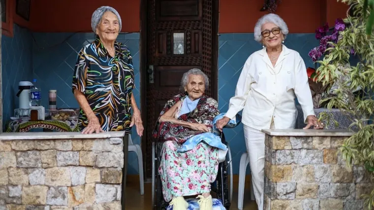 Tres ancianas posan en la entrada de una casa, una sentada en silla de ruedas y dos de pie a sus costados.