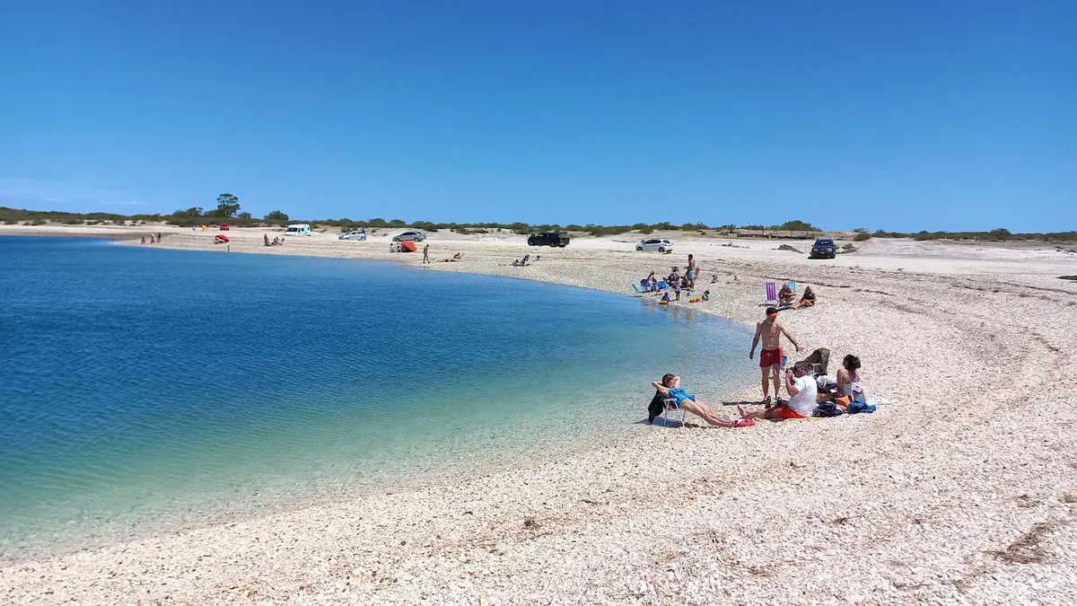 Un grupo de personas descansa en una playa de piedras blancas a orillas de una bahía de aguas calmas con autos estacionados.