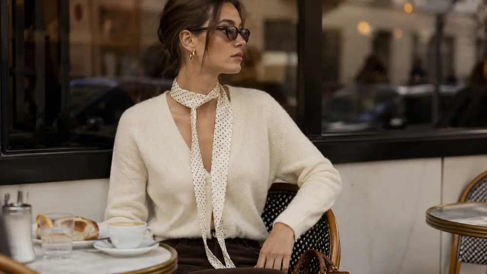 Una mujer con sweater blanco y pañuelo al cuello sentada a la mesa de un bar al aire libre con una taza.