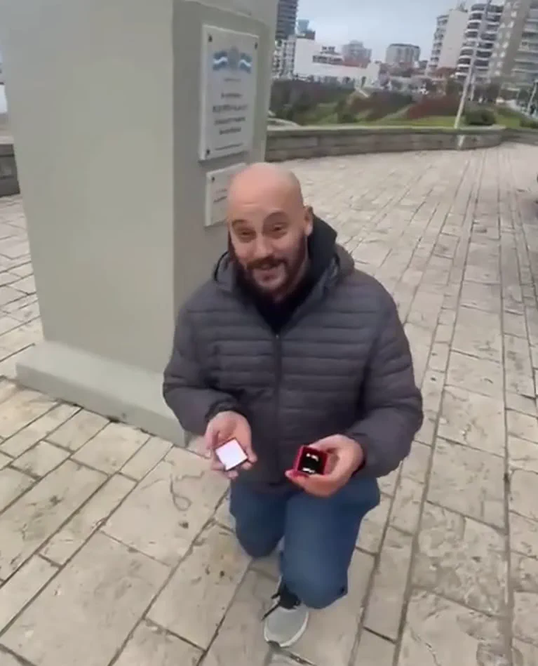 Un hombre arrodillado en una vereda sostiene dos cajitas rojas abiertas con anillos.