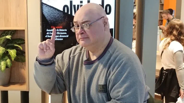 Un hombre calvo con anteojos y buzo gris levanta el dedo índice en un local.