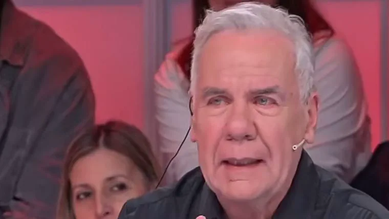 Un hombre de pelo canoso habla con un microfono pegado a la cara en un estudio de television.
