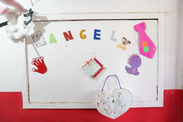 Un panel blanco decorado con letras que forman el nombre Ángel, dibujos infantiles y figuras de goma eva.