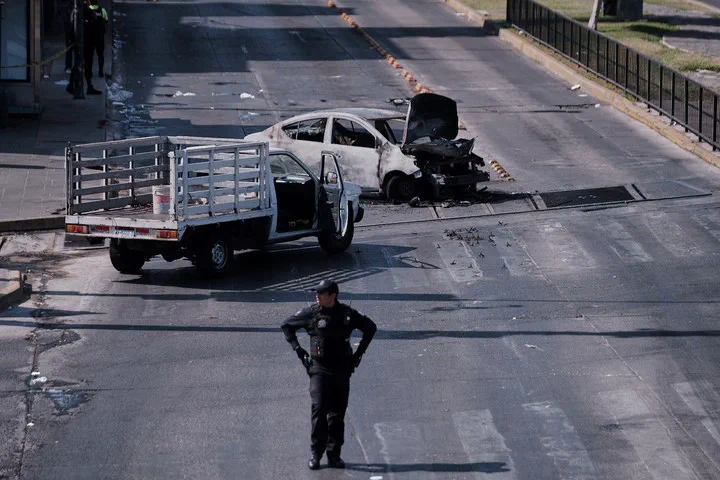 Un policía armado está parado en medio de una avenida donde hay un auto calcinado y una camioneta utilitaria.