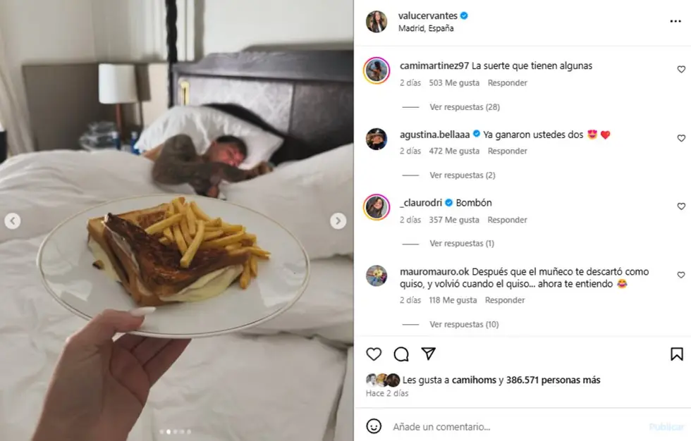 Una mano sostiene un plato con un tostado y papas fritas frente a una cama con una persona acostada.