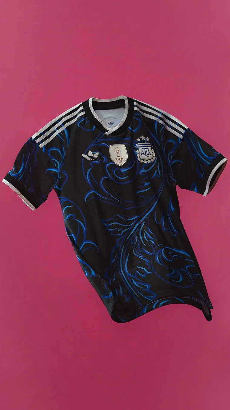 Una camiseta de fútbol negra con vivos azules y el escudo de la AFA está colocada sobre un fondo fucsia.