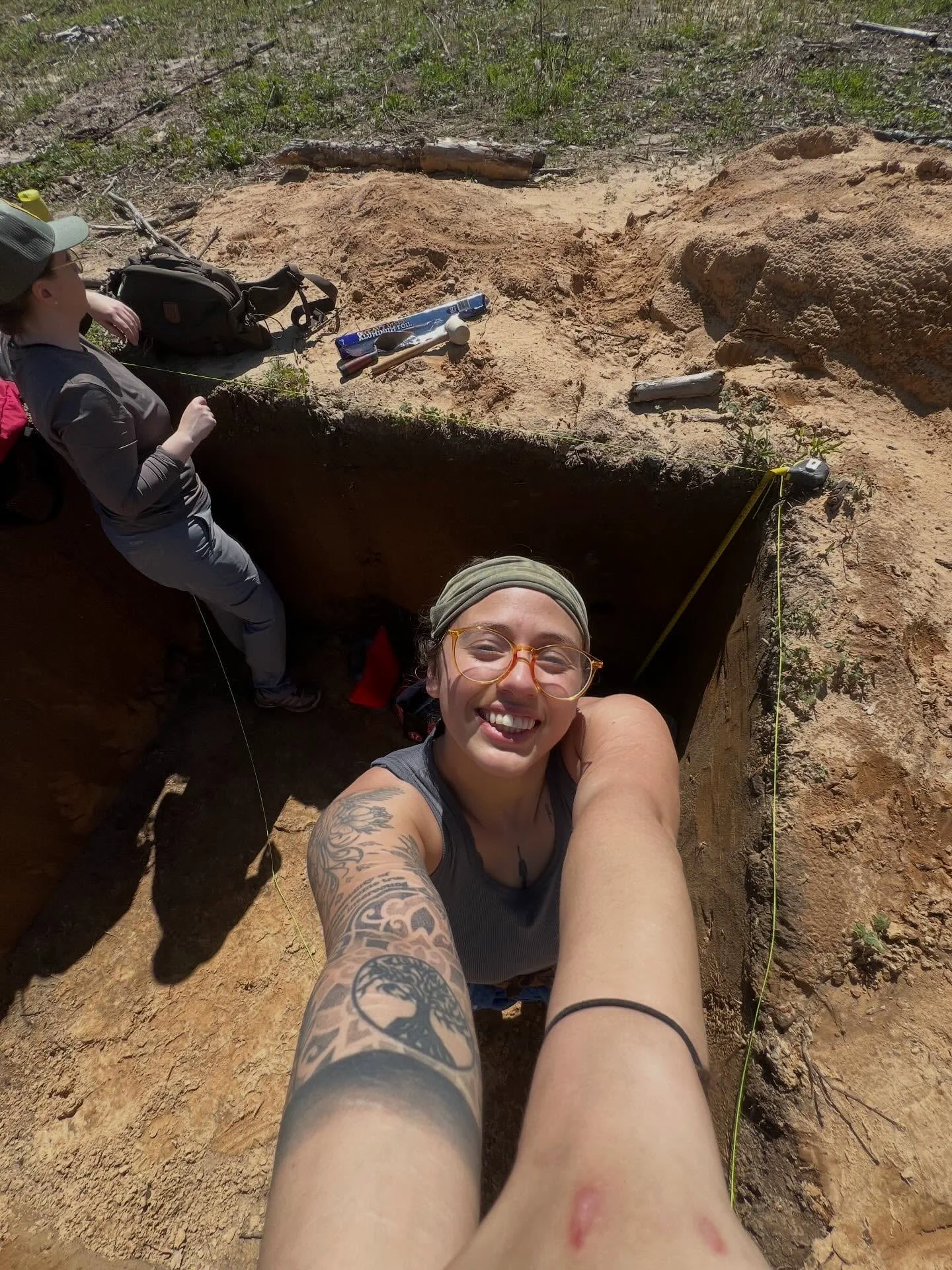 Una chica con anteojos y tatuajes en el brazo se saca una autofoto sonriendo desde el interior de una fosa de tierra.