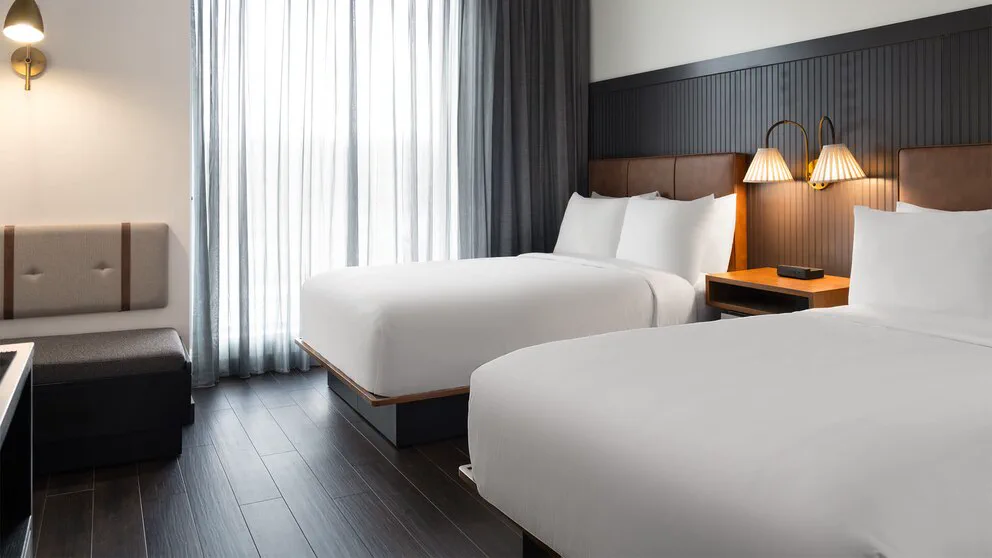 Una habitación de hotel con dos camas individuales hechas, mesas de luz con lámparas y cortinas translúcidas.