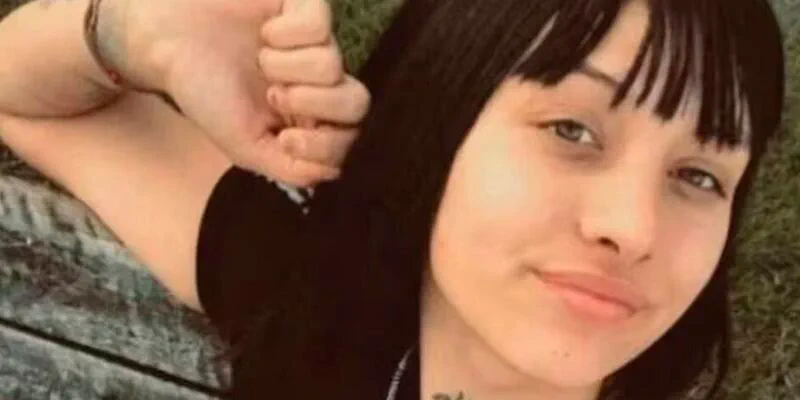 Una joven de flequillo y pelo oscuro sonríe recostada cerca del pasto con un brazo levantado.