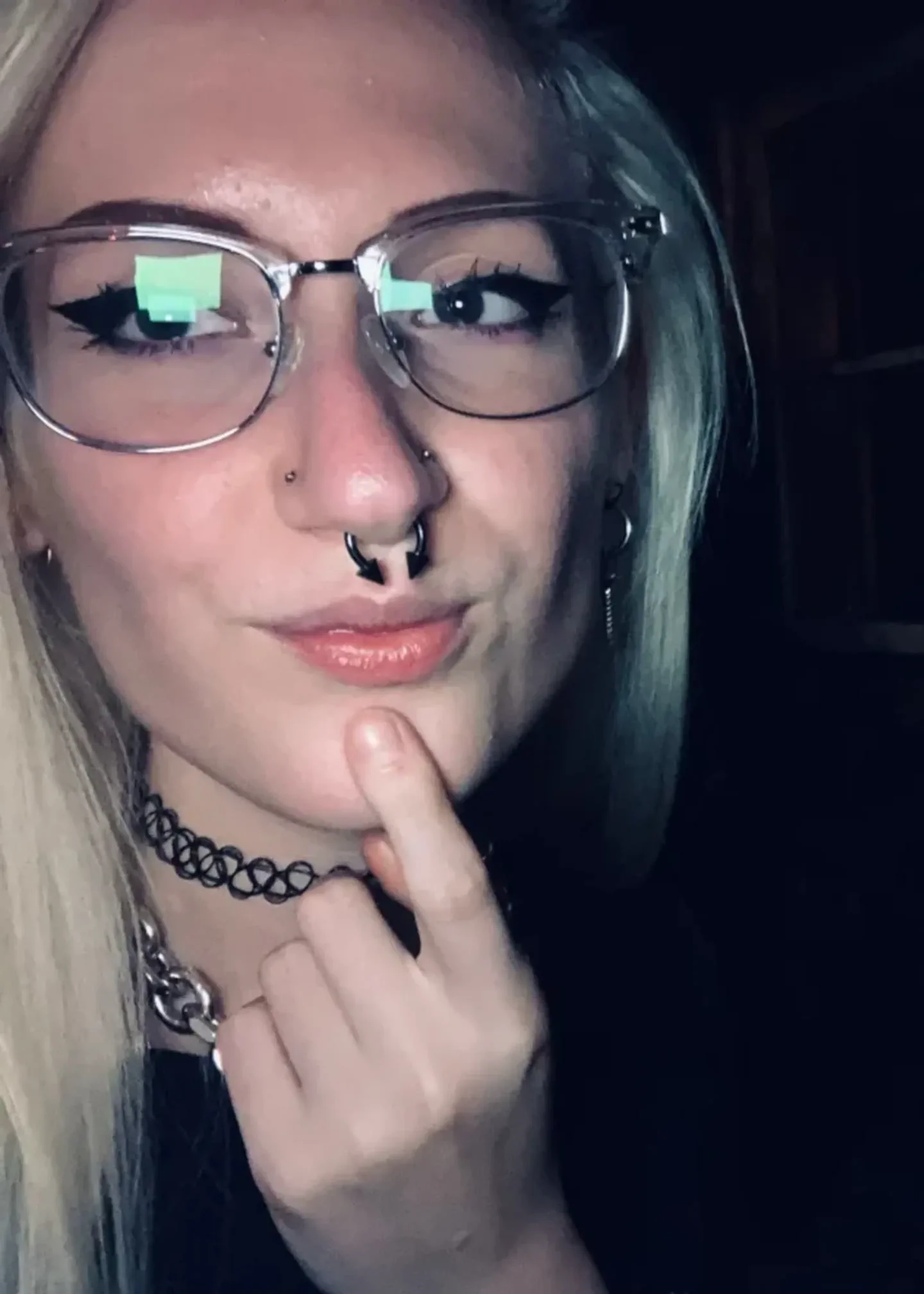 Una joven rubia con anteojos, piercing en la nariz y gargantilla apoya un dedo sobre su menton.