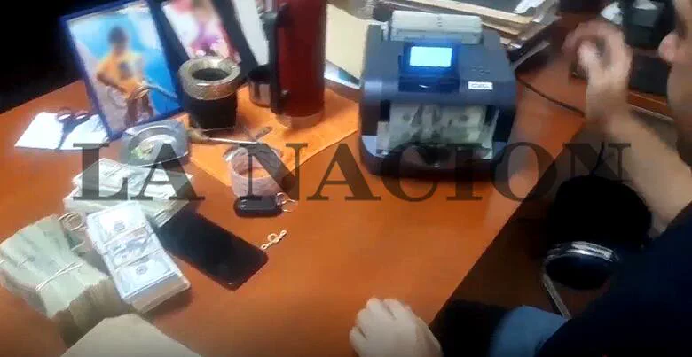 Sobre un escritorio hay fajos de billetes, un celular, un termo, un mate y una máquina contadora de dinero.