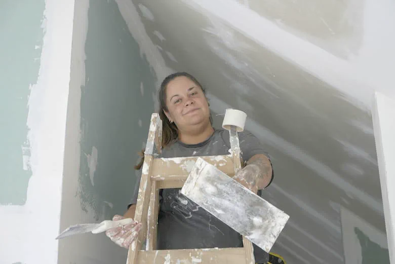 Una mujer subida a una escalera de madera sostiene una espátula y una llana para enduir paredes.