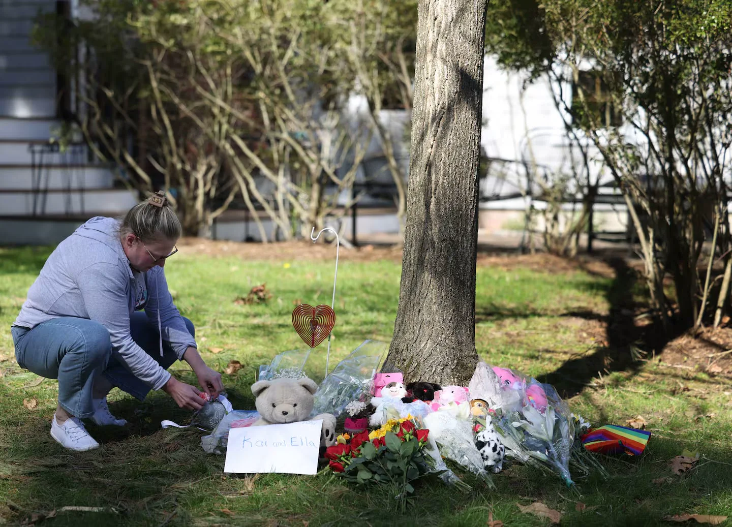 Una mujer se agacha junto a un arbol rodeado de flores, peluches y un cartel con nombres.