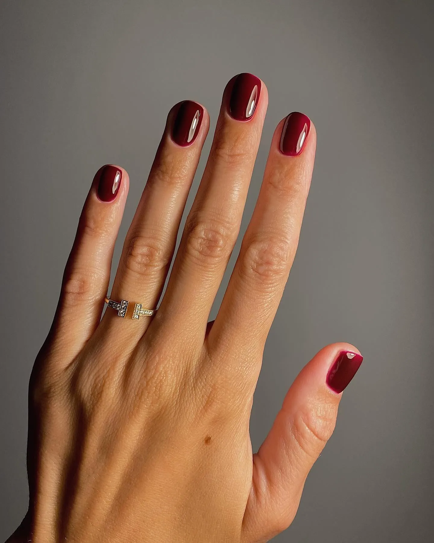 Una mano con las uñas pintadas de bordo y un anillo dorado posa extendida sobre un fondo gris.