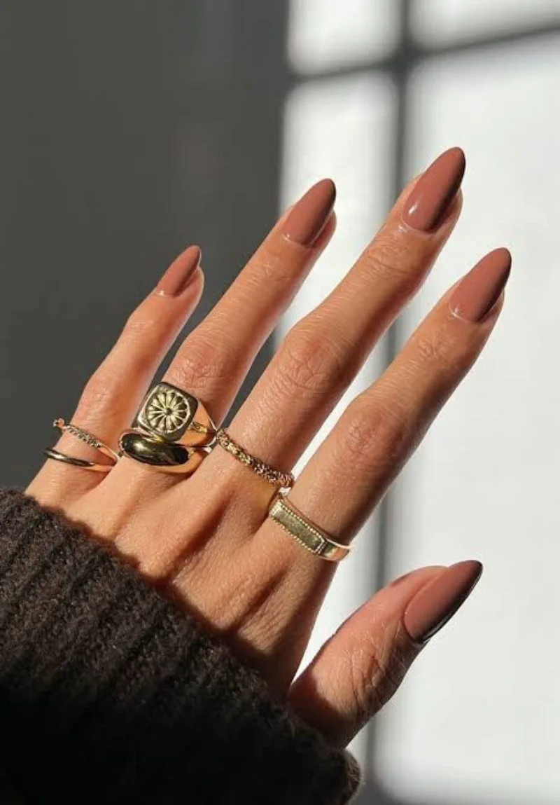 Mano luciendo varios anillos dorados y unas largas pintadas de color marron claro.