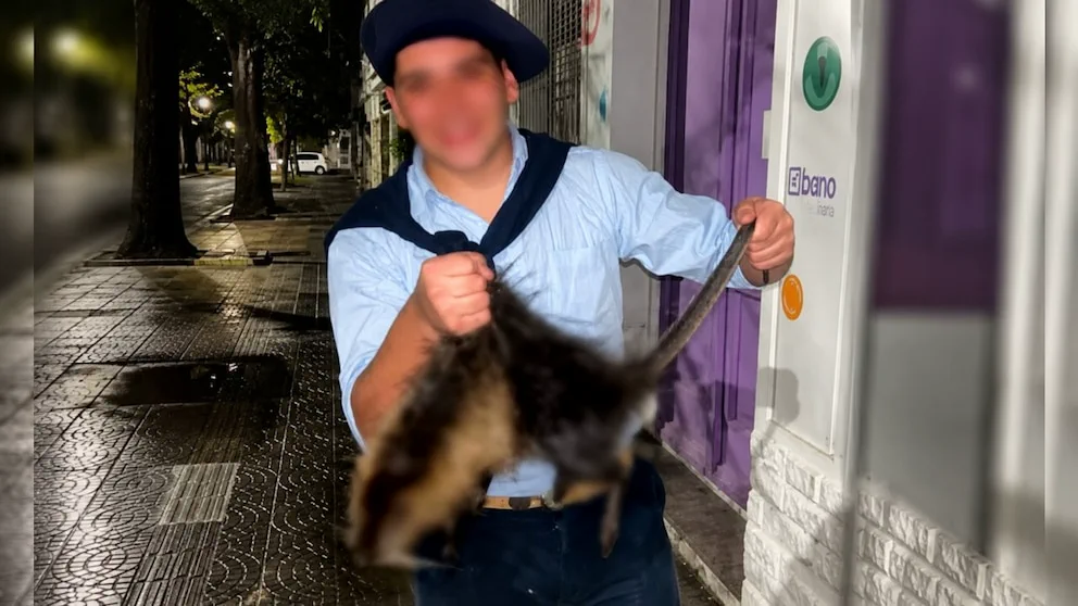 Un joven con boina sosteniendo un coipo muerto en una vereda nocturna frente a un local.