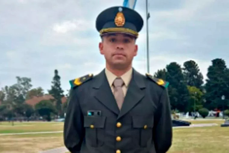 Un militar de uniforme verde y gorra de plato posa de pie al aire libre en un predio verde.