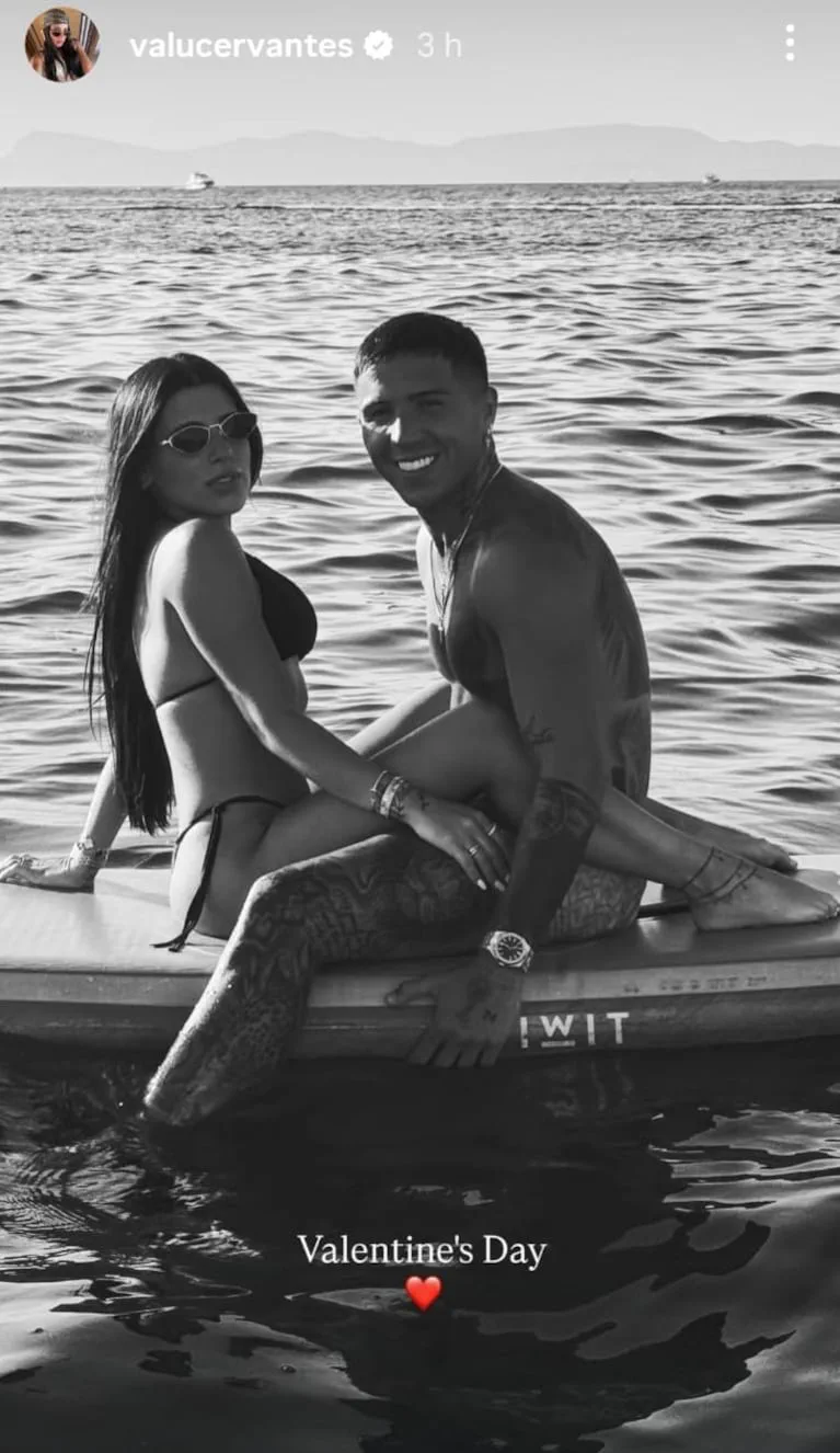 Enzo Fernández y Valentina Cervantes posan sonrientes en traje de baño sentados sobre una tabla en el agua.