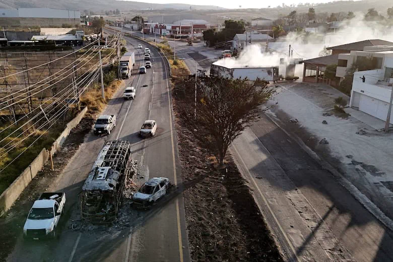 Vehiculos calcinados y un camion humeante bloquean sectores de una avenida con transito y edificaciones a los lados.