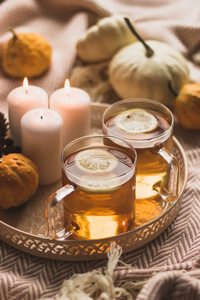 Una bandeja de metal reúne dos tazas de té con rodajas de limón, tres velas encendidas y pequeñas calabazas.