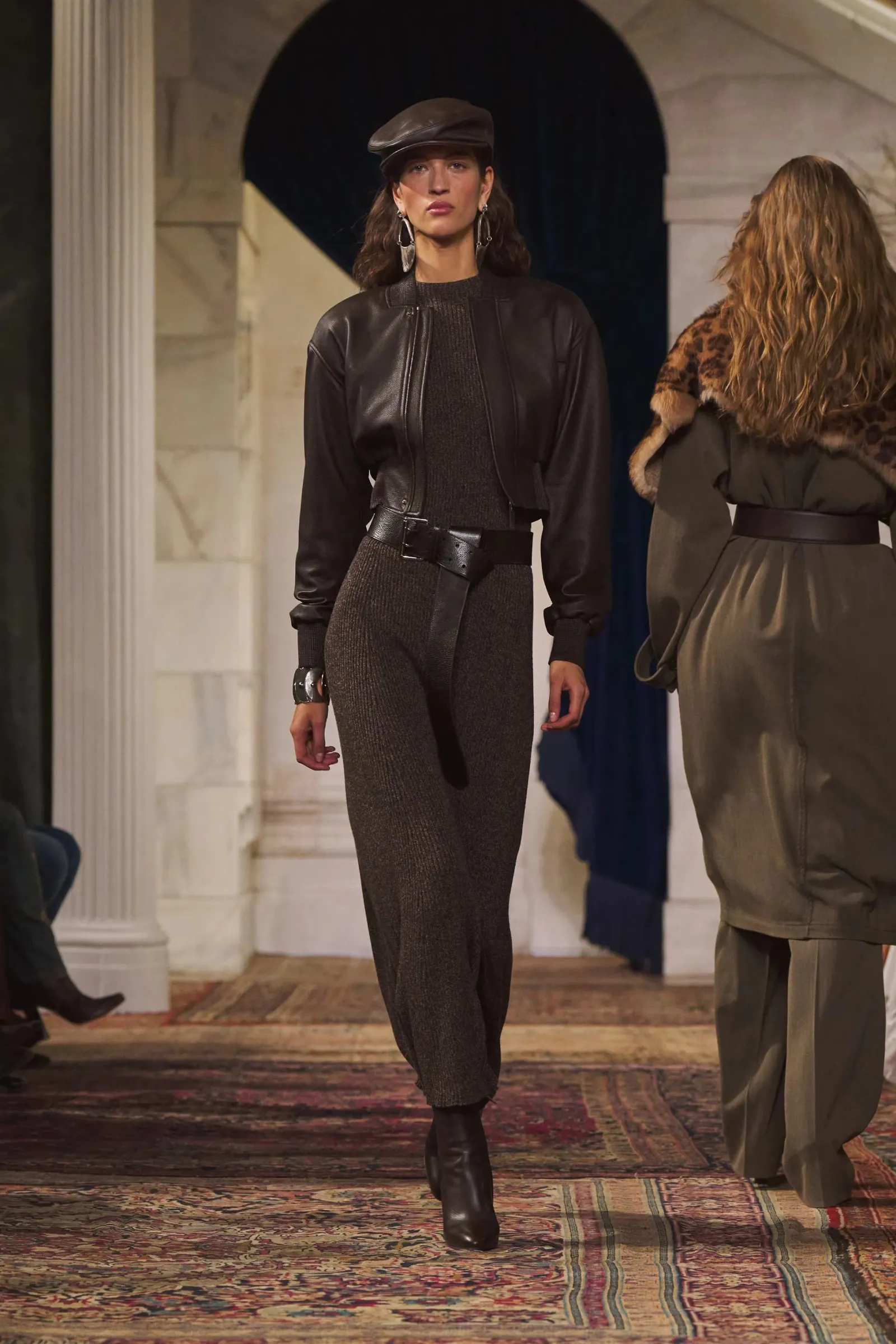Una modelo desfila con vestido tejido marrón, campera corta de cuero, cinturón y boina.