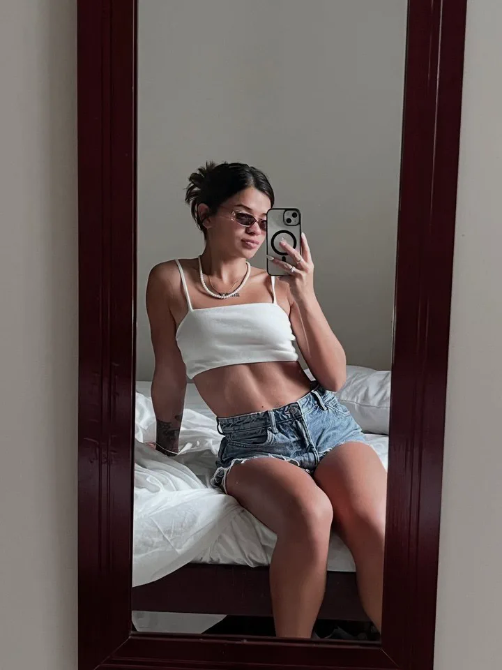 Una chica con anteojos oscuros, top blanco y short de jean se saca una selfie en el espejo sentada en la cama.