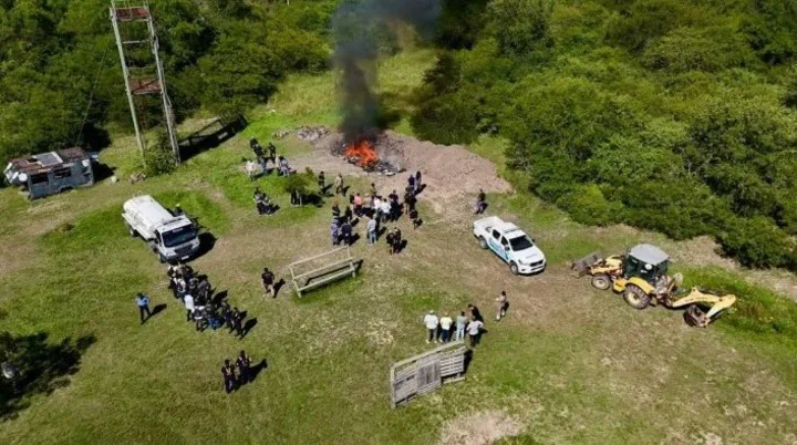 Una toma aérea muestra a personas y vehículos agrupados en un descampado junto a una quema con humo negro.