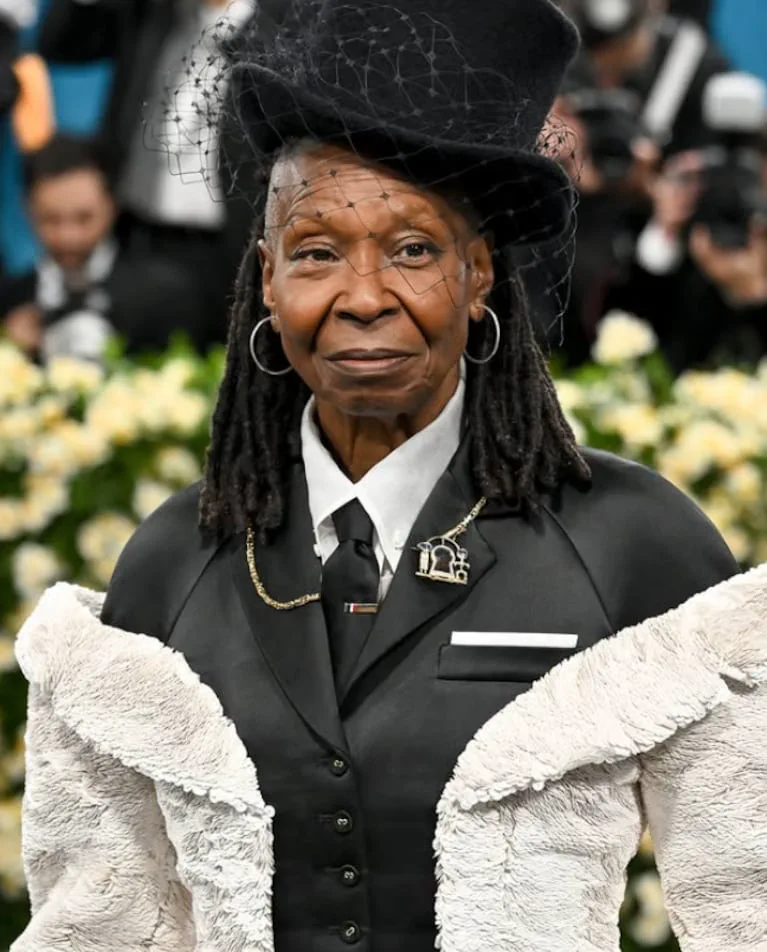 Whoopi Goldberg con galera negra con redecilla, traje oscuro y cuello con piel clara en un evento.