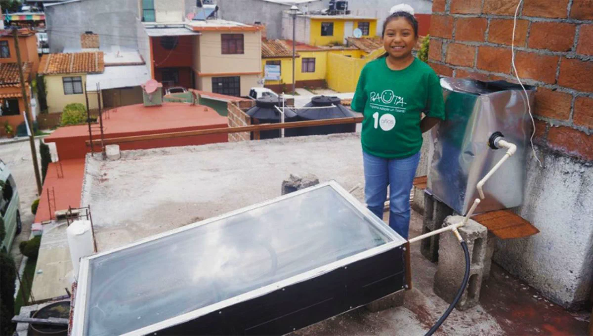 Una nena con remera verde parada en una terraza junto a un calentador solar casero.