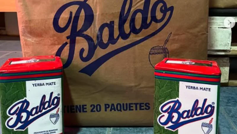 Dos paquetes de yerba mate Baldo apoyados en el suelo delante de una bolsa de papel madera de la misma marca.