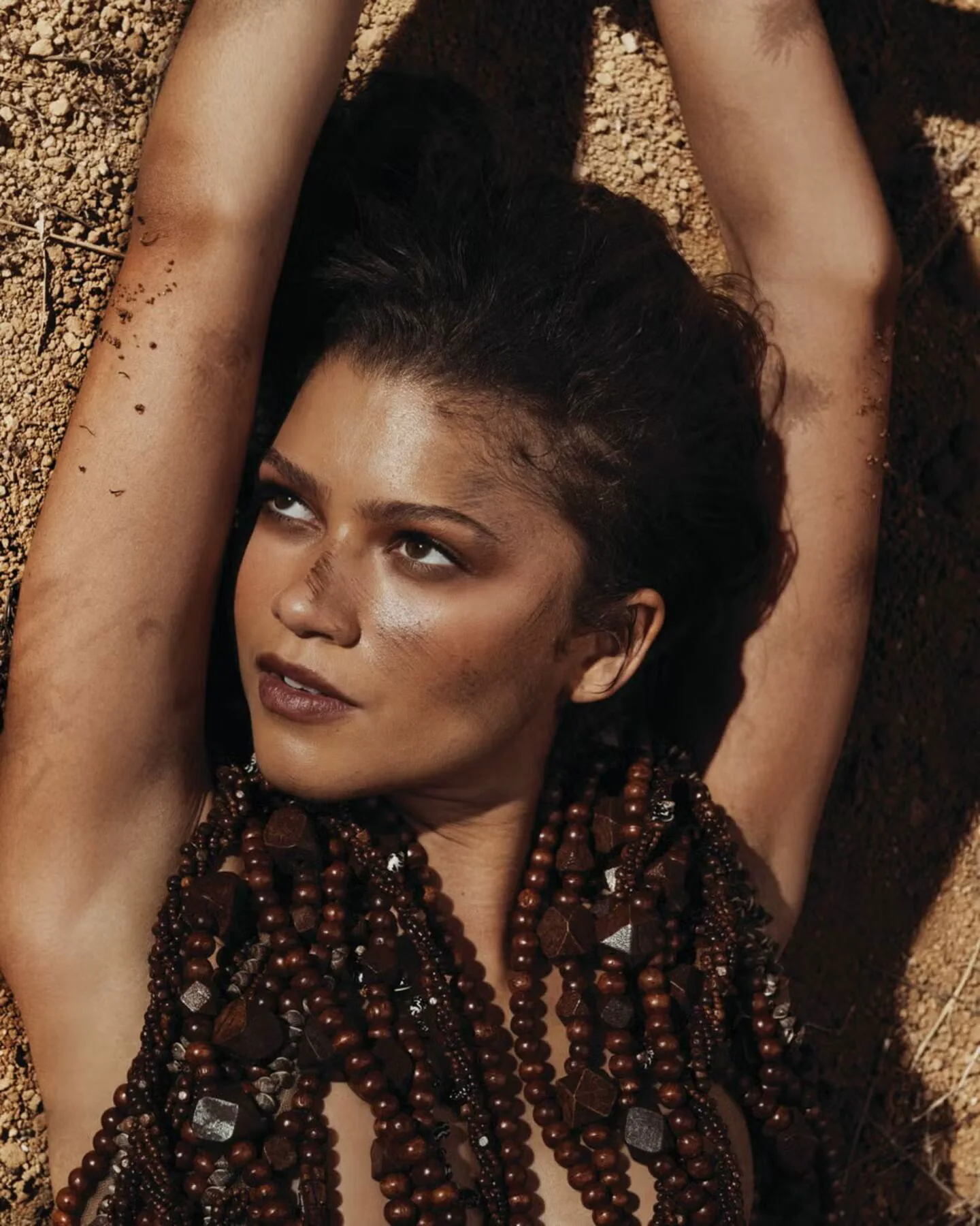 La actriz Zendaya acostada en la tierra con los brazos en alto y collares de cuentas oscuras.
