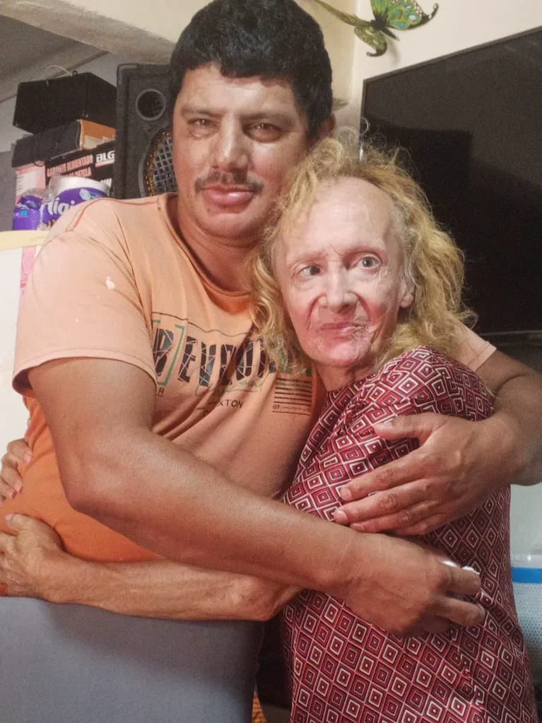 Un hombre con remera naranja abraza a una mujer rubia de contextura delgada vestida con una blusa estampada.