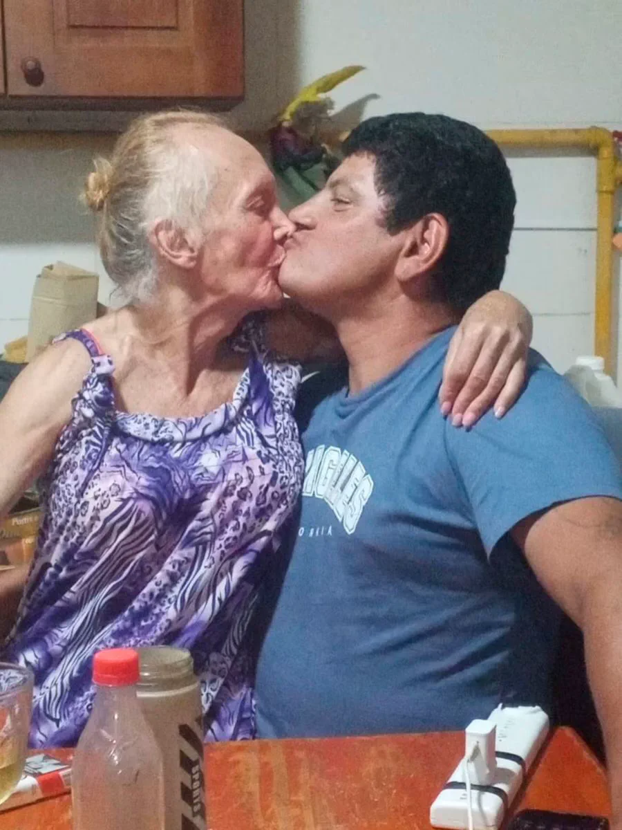 Una mujer mayor y un hombre se dan un beso en la boca sentados a una mesa.
